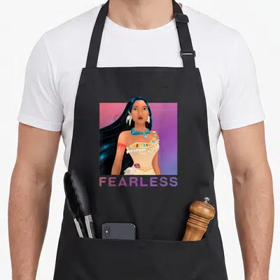 Pocahontas princess apron - FEARLESS text below a cartoon woman portrait in a pink and purple gradient square — shown on a black cotton canvas apron against a white background