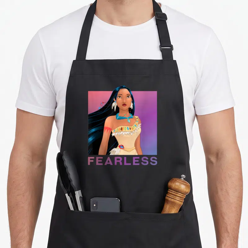 Pocahontas princess apron - FEARLESS text below a cartoon woman portrait in a pink and purple gradient square — shown on a black cotton canvas apron against a white background