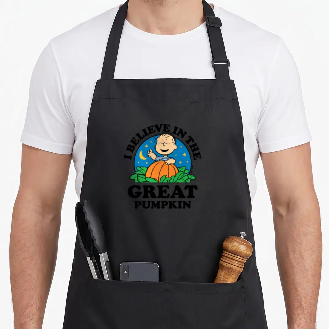 Retro halloween pumpkin apron - Linus leaning on orange pumpkin inside blue circle with I BELIEVE IN THE GREAT PUMPKIN — shown on a black cotton canvas apron against a white background