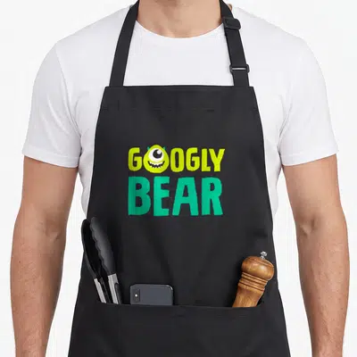 Mike bear apron - GOOGLY BEAR text with monster head replacing O in lime green and teal on black — shown on a black cotton canvas apron against a white background