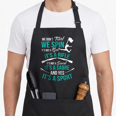 Twirl spin apron - Stacked text WE DON'T Twirl WE SPIN with majorette silhouette and crossed rifles and sabres in white — shown on a black cotton canvas apron against a white background