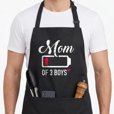 Funny battery apron - Mom OF 3 BOYS text above low red battery icon and heart in distressed white font — shown on a black cotton canvas apron against a white background