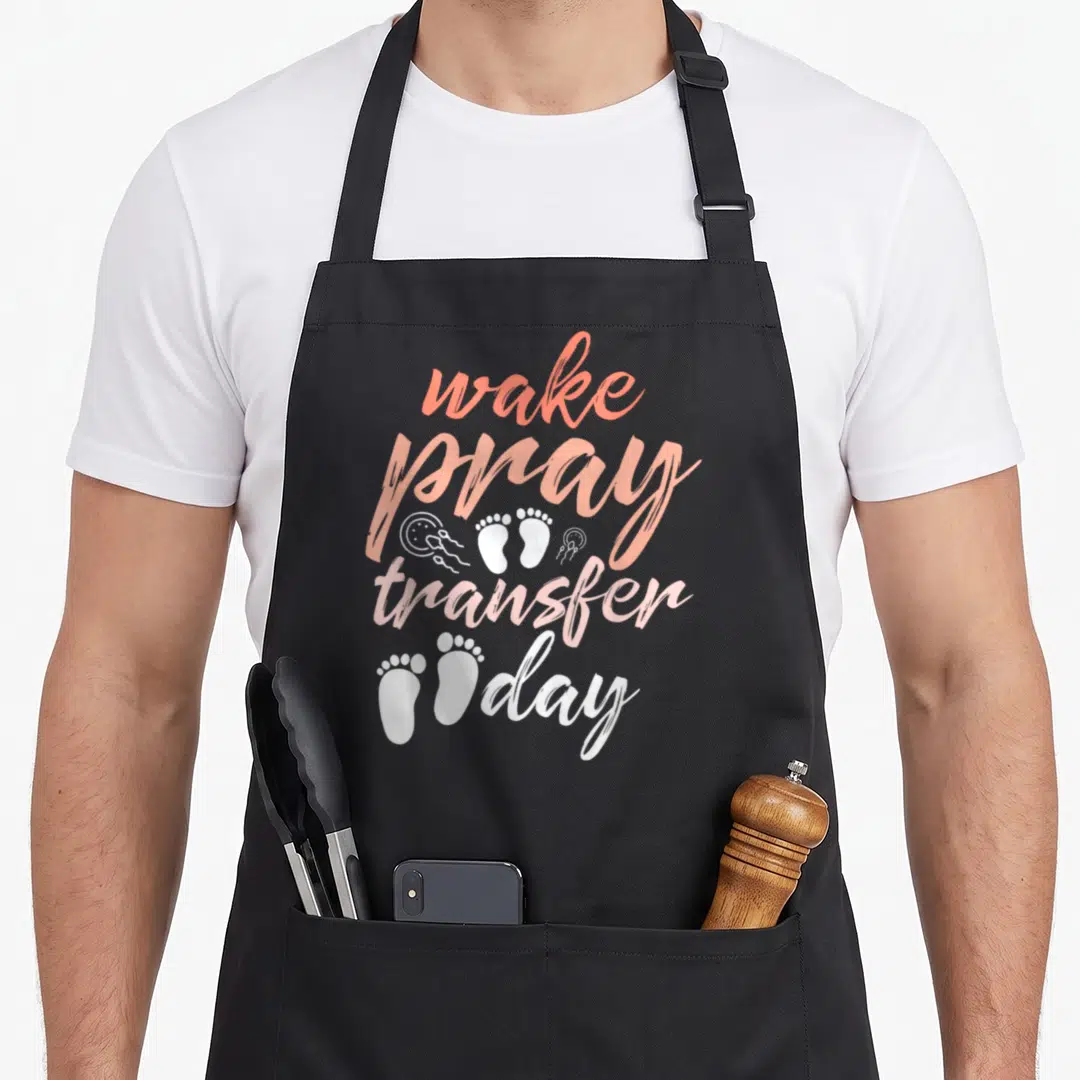 Pray infertility awareness apron - Vertical wake pray transfer day text in coral pink white with footprint and sperm — shown on a black cotton canvas apron against a white background