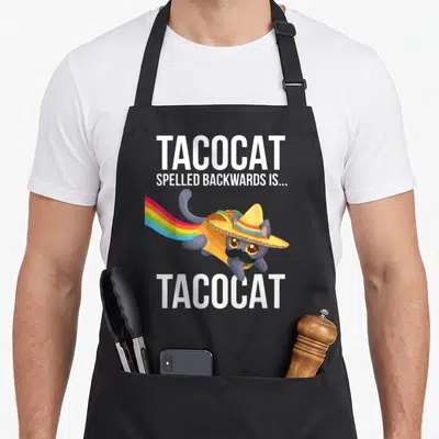 Palindrome taco apron - Cartoon grey cat in sombrero inside taco shell with rainbow tail between TACOCAT text lines — shown on a black cotton canvas apron against a white background