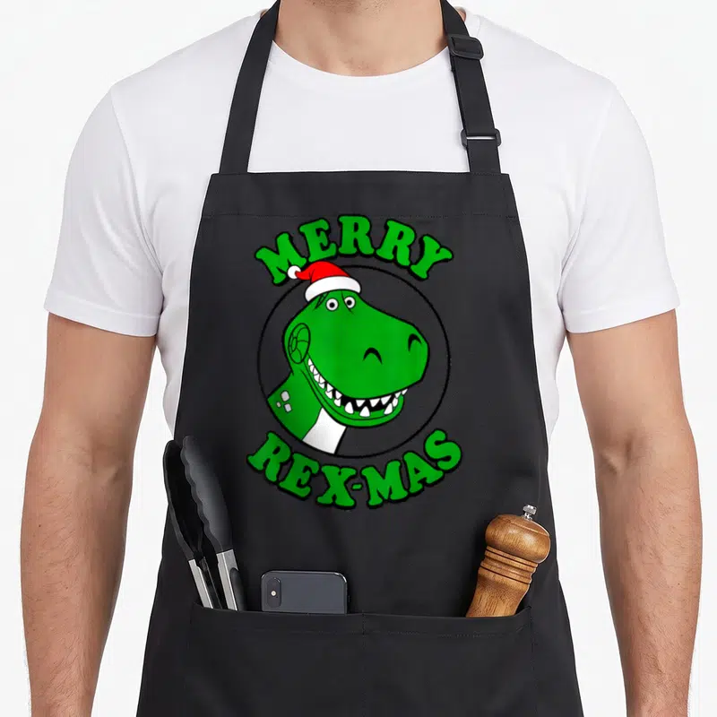 Rex merry christmas apron - Green T-Rex head in black circle with red hat and MERRY REX-MAS text — shown on a black cotton canvas apron against a white background