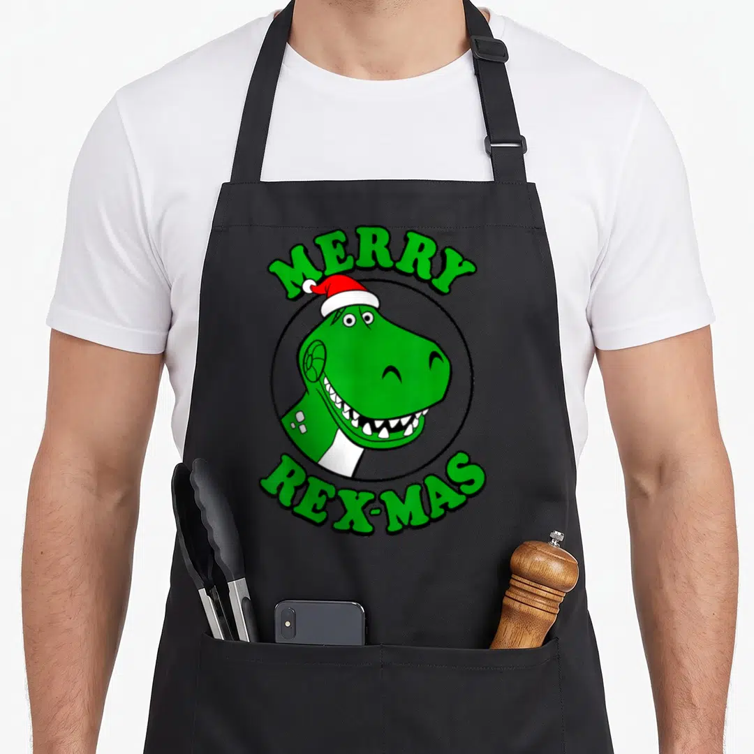 Rex merry christmas apron - Green T-Rex head in black circle with red hat and MERRY REX-MAS text — shown on a black cotton canvas apron against a white background