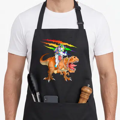 Tyrannosaurus t rex apron - Cartoon white unicorn with rainbow mane riding orange T-Rex with blue sunglasses — shown on a black cotton canvas apron against a white background
