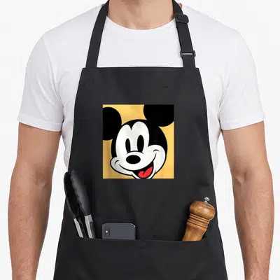 Boxed essential apron - Mickey Mouse head centered on yellow square with black ears and red tongue — shown on a black cotton canvas apron against a white background