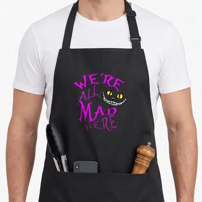Mad cat apron - Cheshire Cat face with We're All Mad Here text in pink purple yellow white — shown on a black cotton canvas apron against a white background