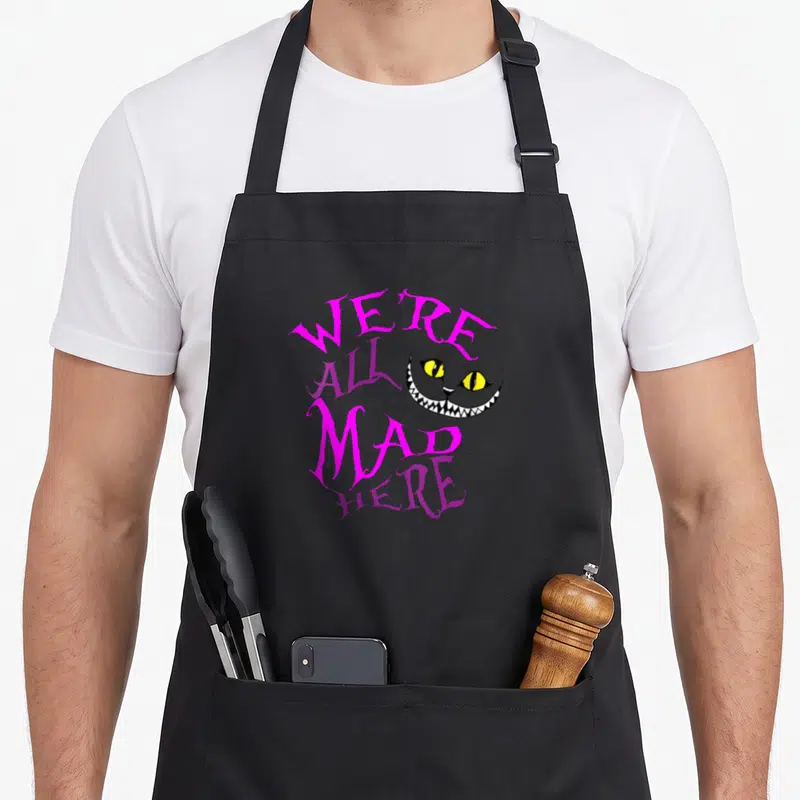 Mad cat apron - Cheshire Cat face with We're All Mad Here text in pink purple yellow white — shown on a black cotton canvas apron against a white background