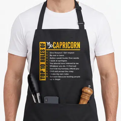 Rule capricorn birthday apron - Yellow and white typography listing ten Capricorn rules including sarcasm and respect — shown on a black cotton canvas apron against a white background