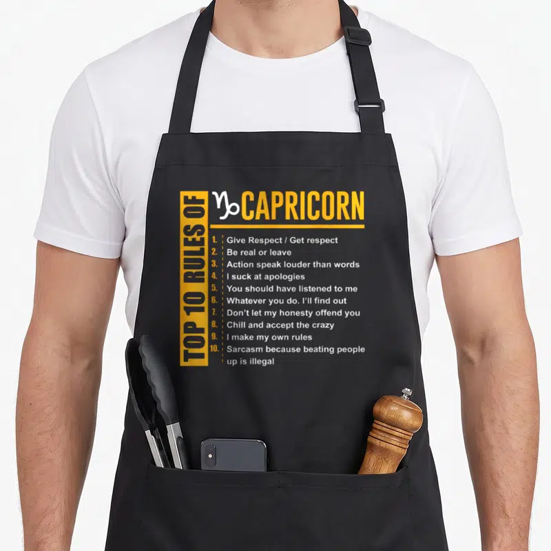 Rule capricorn birthday apron - Yellow and white typography listing ten Capricorn rules including sarcasm and respect — shown on a black cotton canvas apron against a white background