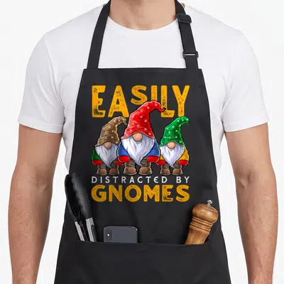 Easily gnome apron - Three cartoon gnomes in red, green, and brown hats below yellow text reading EASILY DISTRACTED — shown on a black cotton canvas apron against a white background