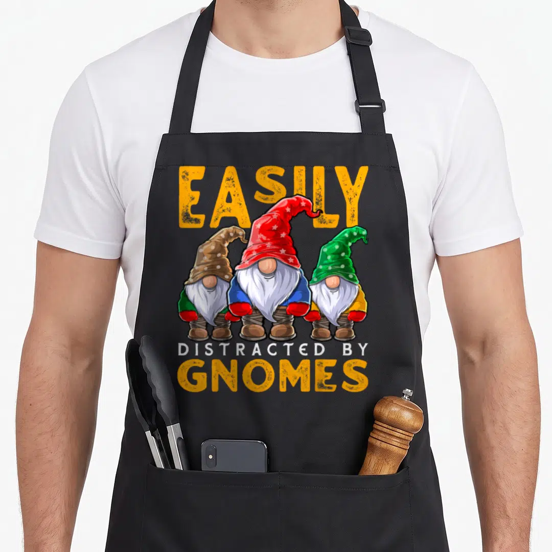 Easily gnome apron - Three cartoon gnomes in red, green, and brown hats below yellow text reading EASILY DISTRACTED — shown on a black cotton canvas apron against a white background