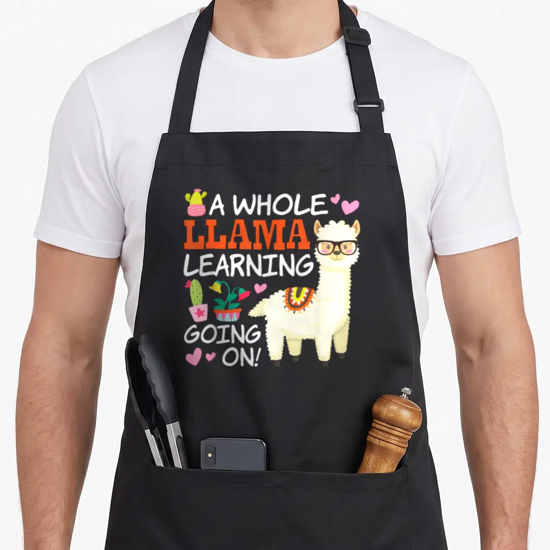 Llama teacher apron - Cartoon llama with glasses and stacked text A WHOLE LLAMA LEARNING GOING ON surrounded by cacti — shown on a black cotton canvas apron against a white background