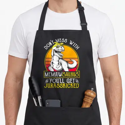 Jurasskicked grandma apron - T-Rex with sunglasses in front of striped sun with text Don't Mess With Memawsaurus — shown on a black cotton canvas apron against a white background