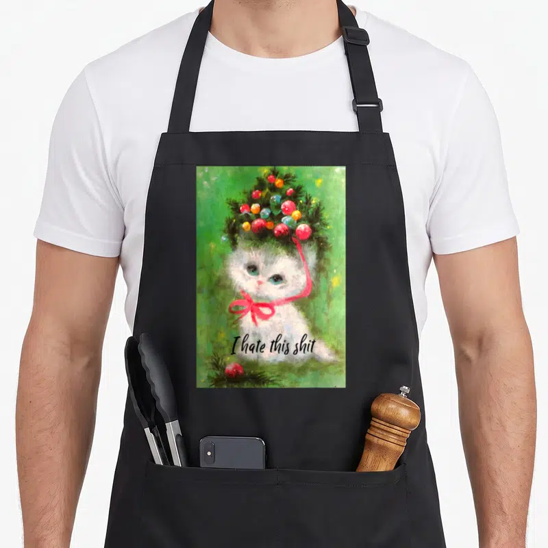 Kitsch crazy cat lady apron - Watercolor kitten with Christmas tree hat and 'I hate this shit' text on black apron — shown on a black cotton canvas apron against a white background