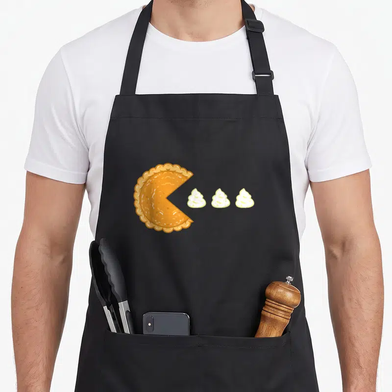 Gamer pie apron - Orange pumpkin pie with missing slice and three white whipped cream dollops in cartoon style — shown on a black cotton canvas apron against a white background