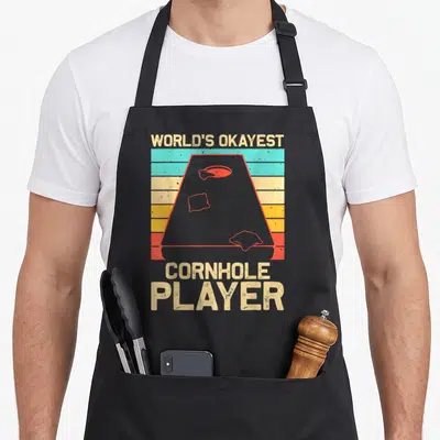 Game bags and aprons - Retro cornhole board on striped background with WORLD'S OKAYEST CORNHOLE PLAYER text — shown on a black cotton canvas apron against a white background