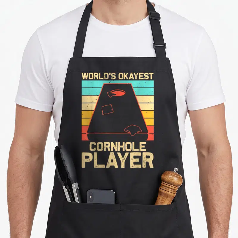 Game bags and aprons - Retro cornhole board on striped background with WORLD'S OKAYEST CORNHOLE PLAYER text — shown on a black cotton canvas apron against a white background