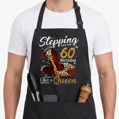 Heel 60th birthday apron - Leopard high heels with crown, red lips, and 60th birthday text in gold and white — shown on a black cotton canvas apron against a white background