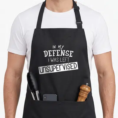 Defense unsupervised remaining apron - White stacked text reading In My Defense I Was Left Unsupervised on a grey — shown on a black cotton canvas apron against a white background