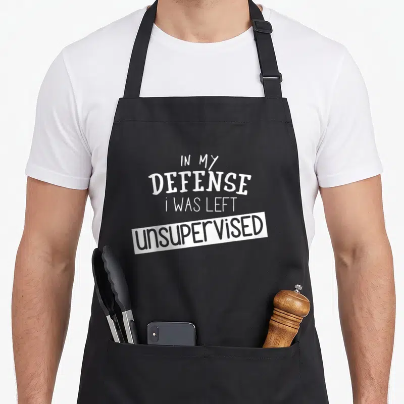 Defense unsupervised remaining apron - White stacked text reading In My Defense I Was Left Unsupervised on a grey — shown on a black cotton canvas apron against a white background