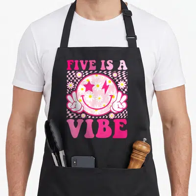 Five custom face apron - Pink daisy smiley face with peace hands and FIVE IS A VIBE text on checkerboard background — shown on a black cotton canvas apron against a white background