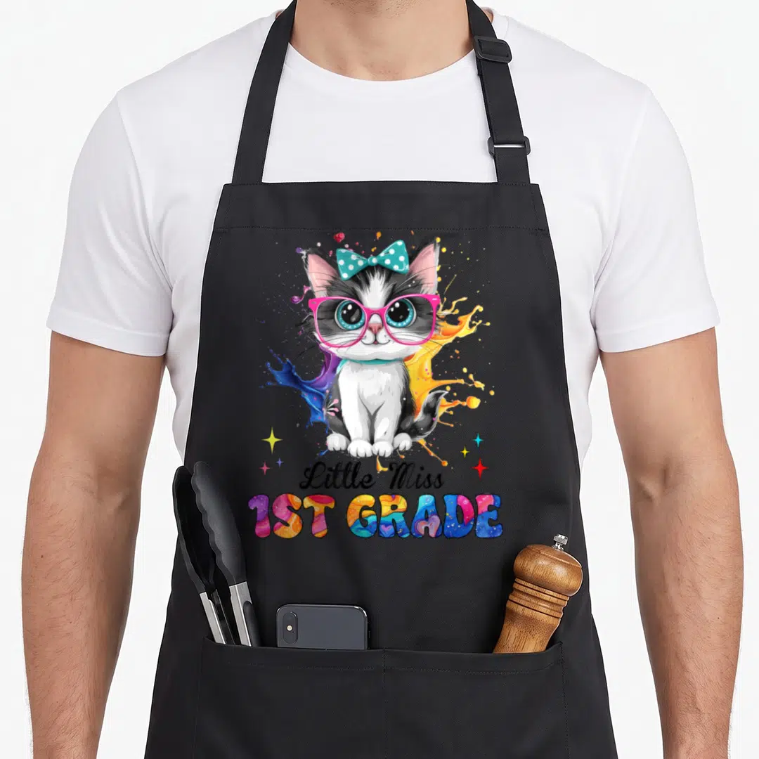 Miss cat apron - Cartoon kitten with pink glasses and teal bow over paint splashes, text Little Miss 1ST GRADE — shown on a black cotton canvas apron against a white background