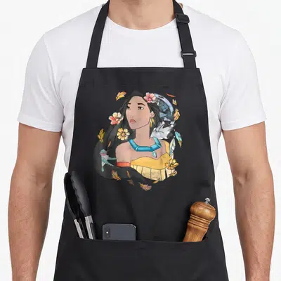 Pocahontas pocatello apron - Watercolor woman with blue necklace, hummingbird, raccoon, and scattered orange and yellow — shown on a black cotton canvas apron against a white background