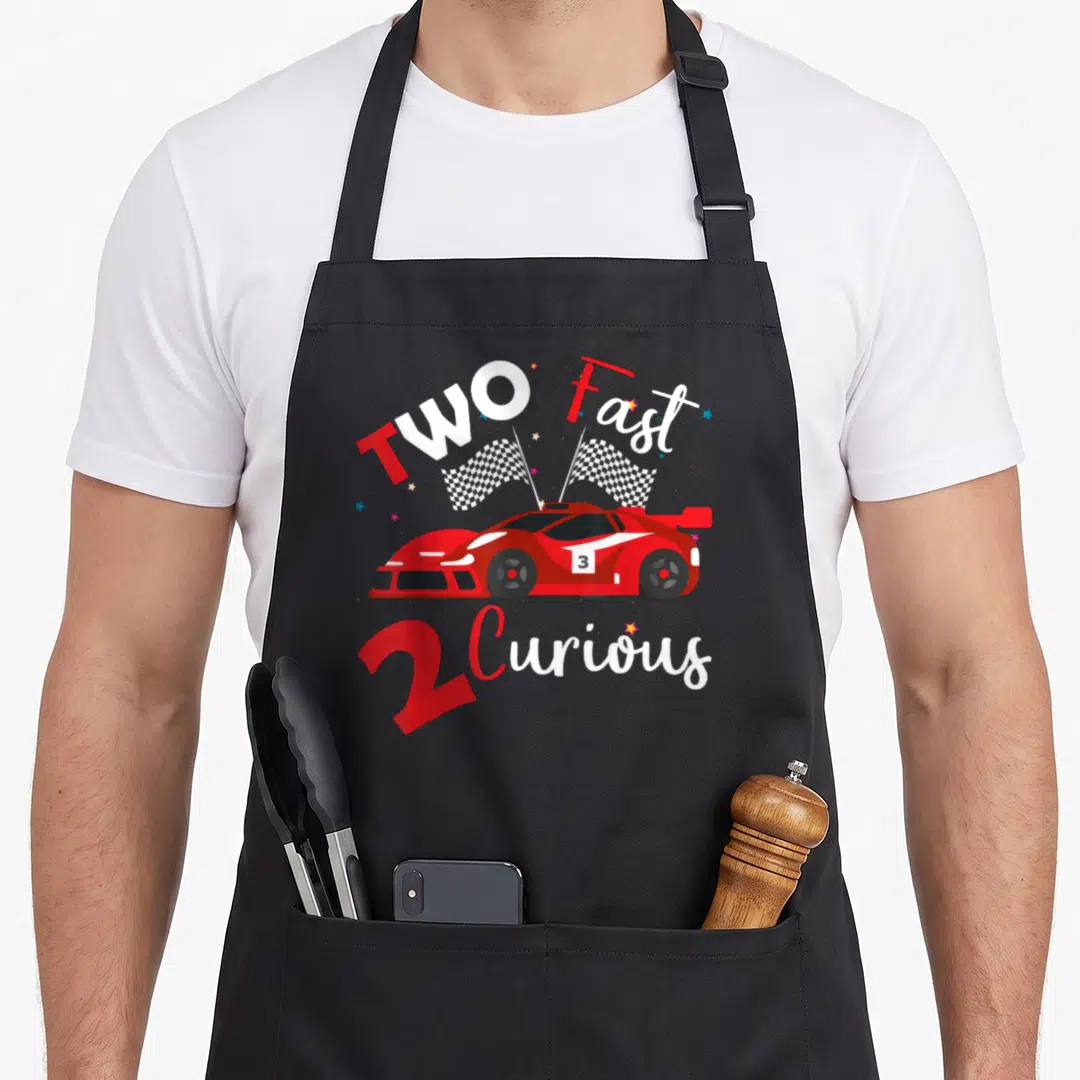 Curious racing apron - Red cartoon race car with checkered flags and TWO Fast 2 Curious text — shown on a black cotton canvas apron against a white background