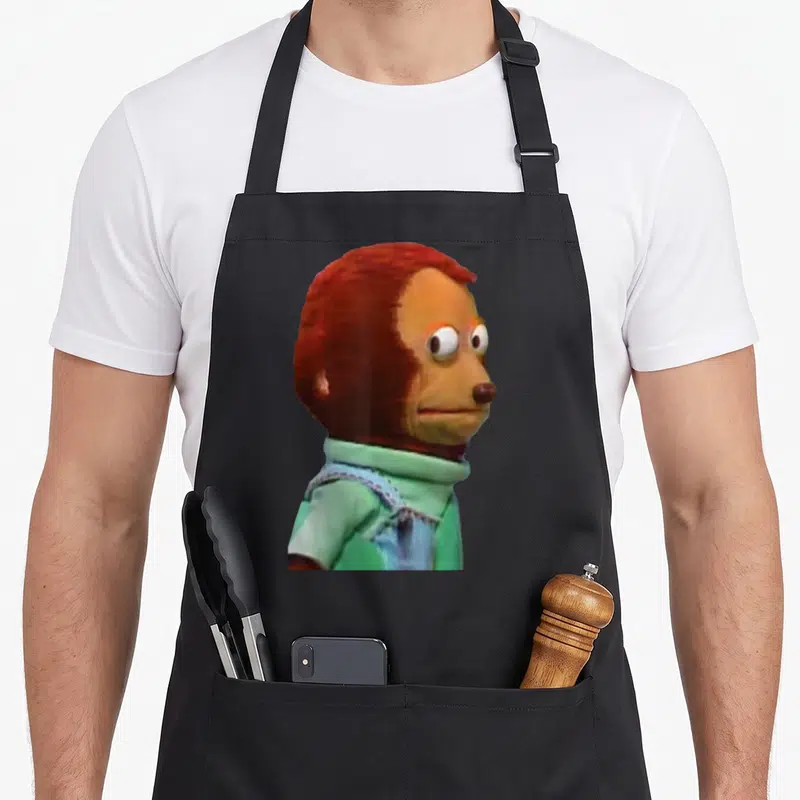 Awkward doll apron - Brown monkey puppet in green shirt facing right with wide eyes — shown on a black cotton canvas apron against a white background