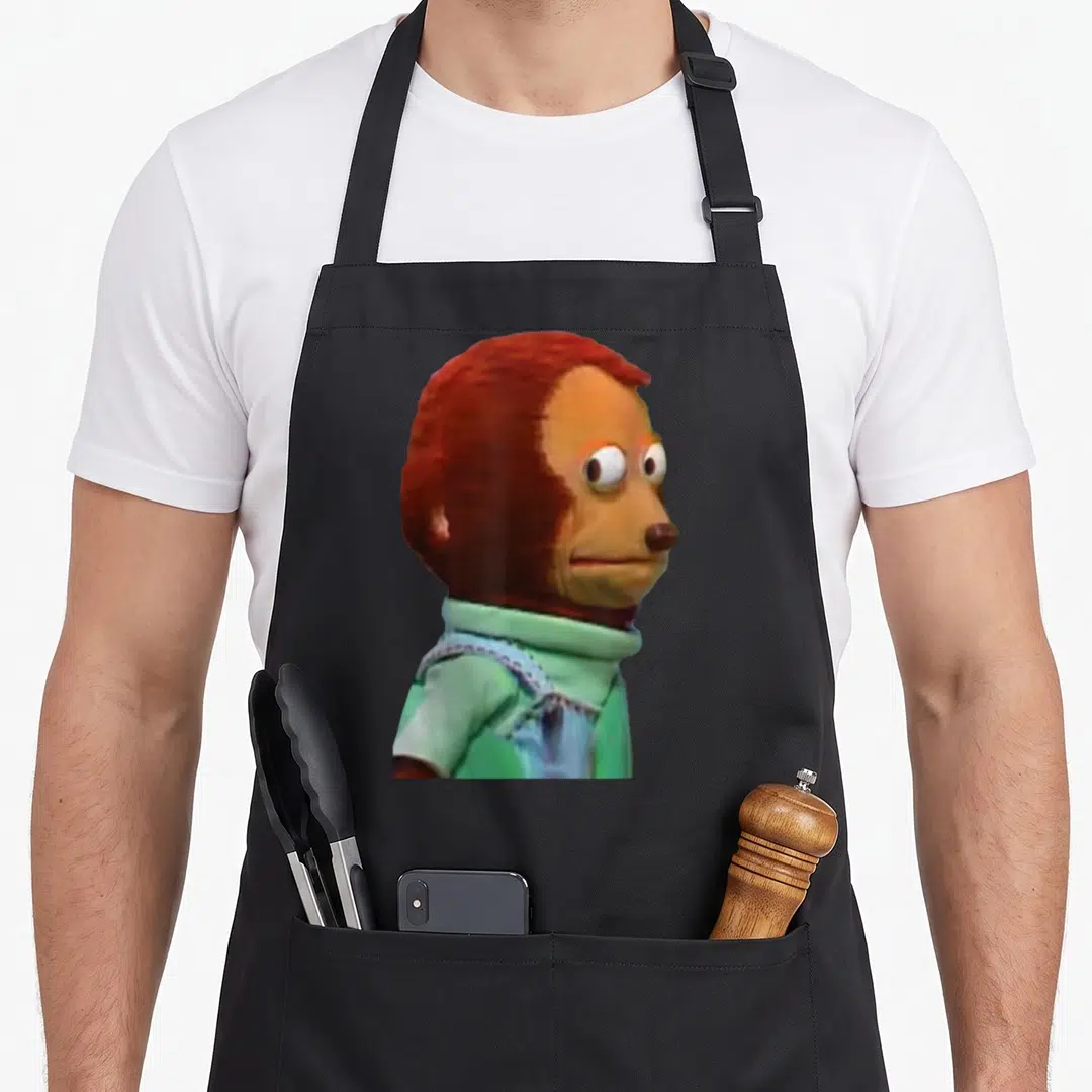 Awkward doll apron - Brown monkey puppet in green shirt facing right with wide eyes — shown on a black cotton canvas apron against a white background