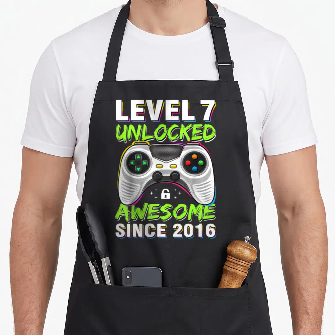 7th gaming aprons - Game controller with Level 7 Unlocked text and lock icon in white lime green and black — shown on a black cotton canvas apron against a white background
