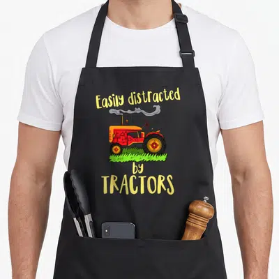 Easily tractor girl apron - Cartoon red tractor on green grass with smoke and yellow text reading Easily distracted — shown on a black cotton canvas apron against a white background