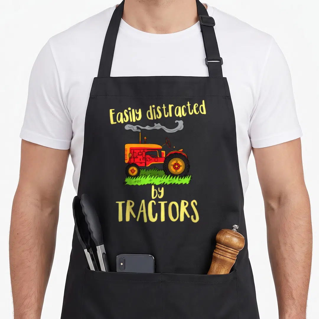 Easily tractor girl apron - Cartoon red tractor on green grass with smoke and yellow text reading Easily distracted — shown on a black cotton canvas apron against a white background