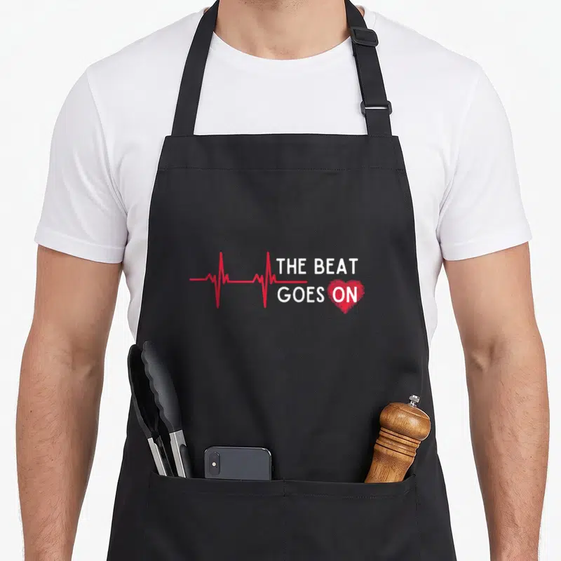 Attack heartbeat funny apron - Red heartbeat line and white text THE BEAT GOES ON ending in a red heart on black — shown on a black cotton canvas apron against a white background