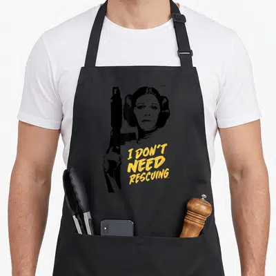 Escape novelty apron - Black silhouette of a woman holding a gun beside yellow text reading I DON'T NEED RESCUING — shown on a black cotton canvas apron against a white background