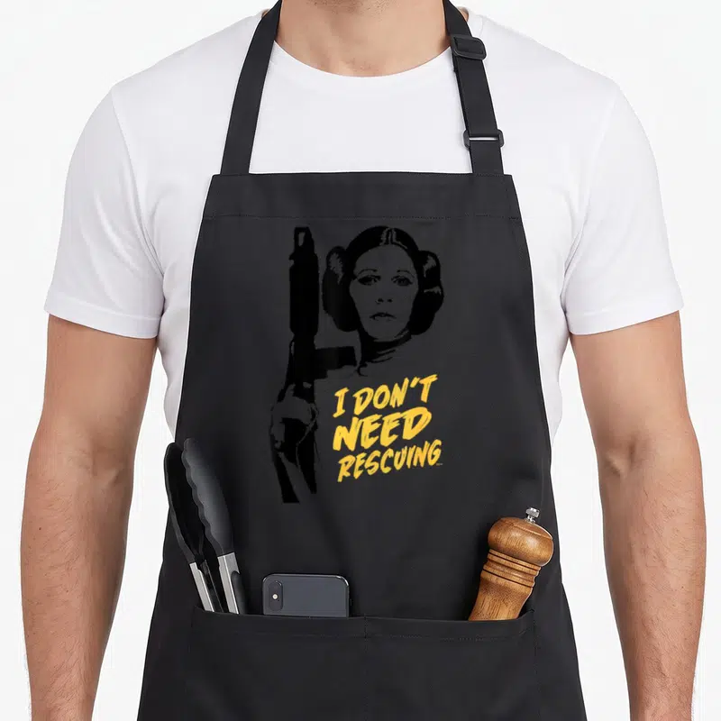 Escape novelty apron - Black silhouette of a woman holding a gun beside yellow text reading I DON'T NEED RESCUING — shown on a black cotton canvas apron against a white background