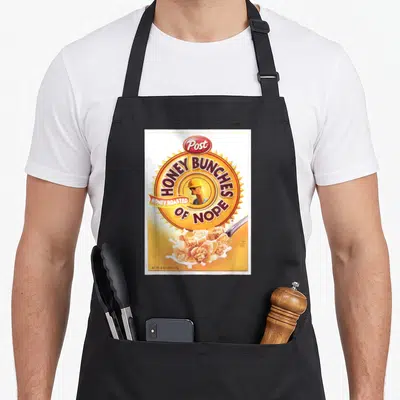 Honey breakfast apron - Cereal box parody with Post HONEY BUNCHES OF NOPE text and man in hard hat — shown on a black cotton canvas apron against a white background