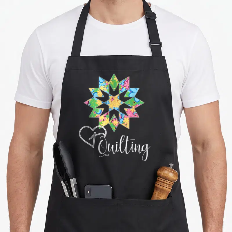 Sewing crafter apron - Multicolored floral quilt block above white heart outline and Quilting script — shown on a black cotton canvas apron against a white background