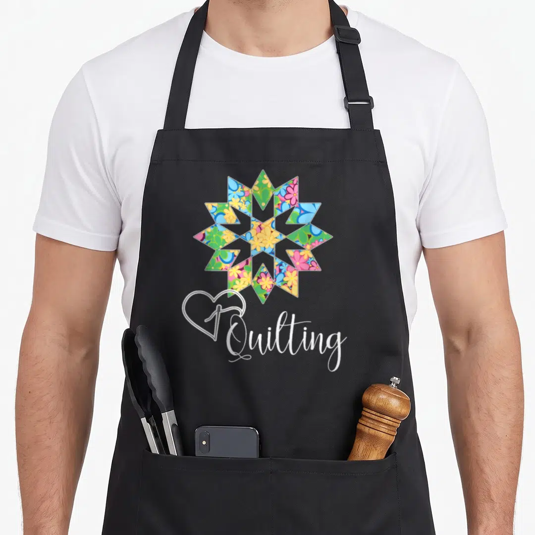 Sewing crafter apron - Multicolored floral quilt block above white heart outline and Quilting script — shown on a black cotton canvas apron against a white background