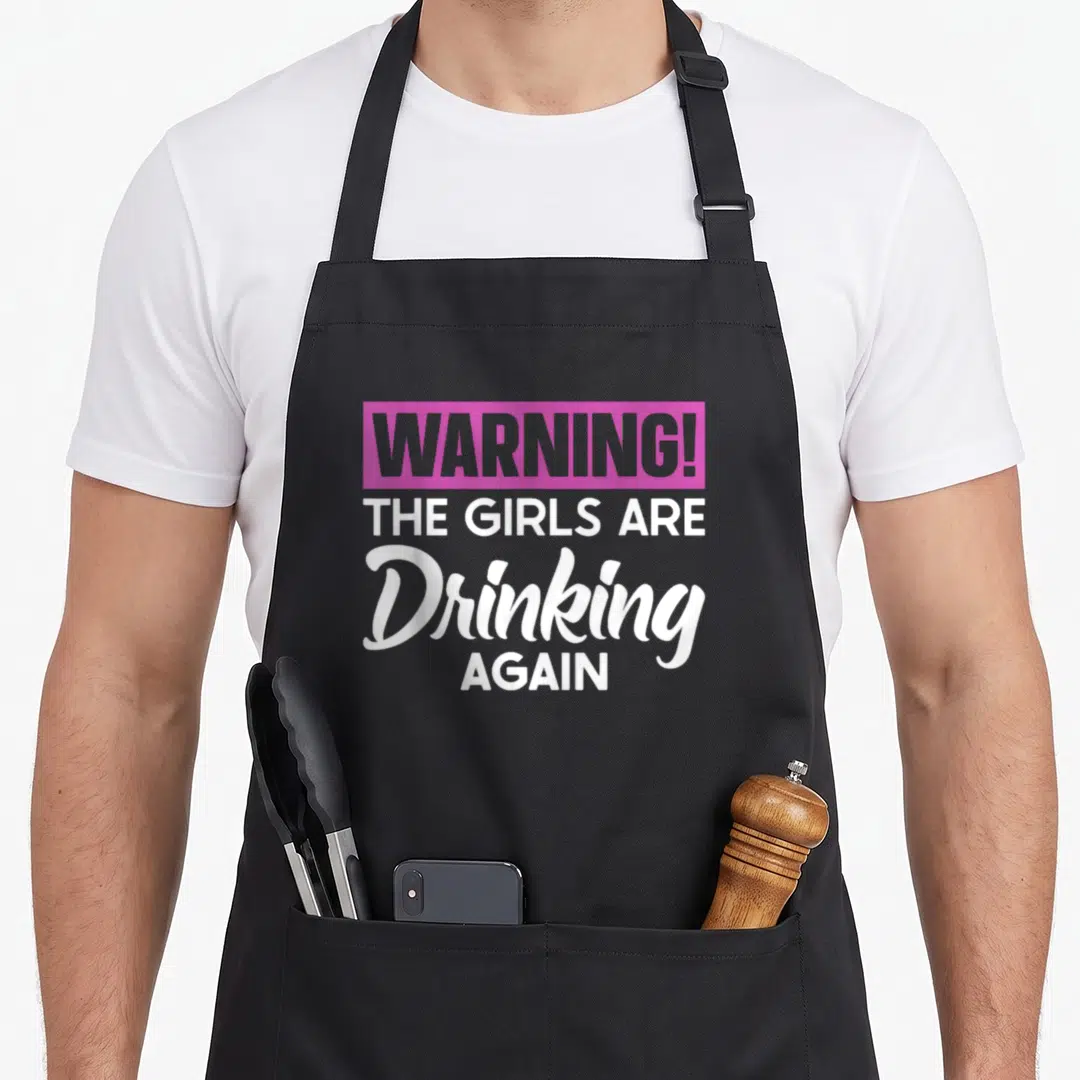 Warning novelty apron - Pink block with WARNING text above white stacked letters reading THE GIRLS ARE Drinking AGAIN — shown on a black cotton canvas apron against a white background