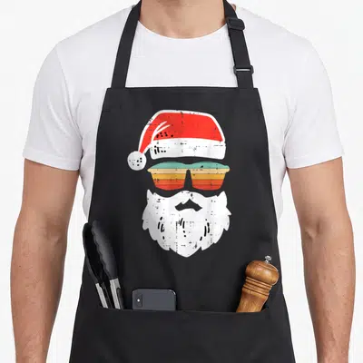 Face christmas santa apron - Retro Santa hat, teal orange sunglasses, and white beard stack — shown on a black cotton canvas apron against a white background