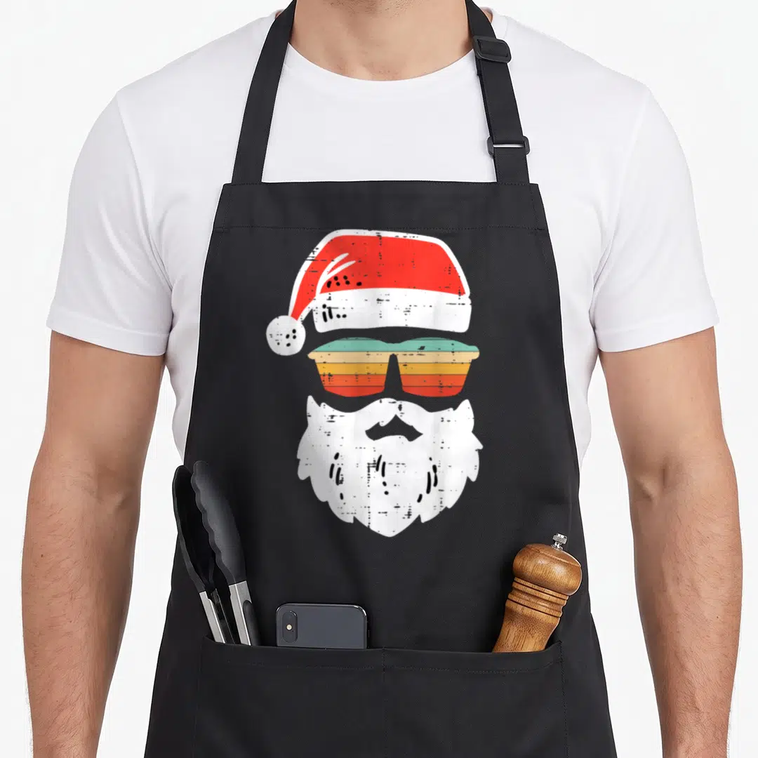 Face christmas santa apron - Retro Santa hat, teal orange sunglasses, and white beard stack — shown on a black cotton canvas apron against a white background