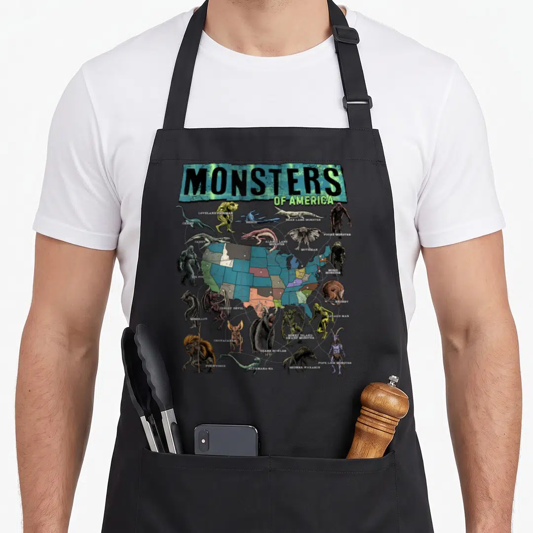 Cryptid folklore apron - Monsters of America map with illustrated cryptids like Big Foot, Mothman, and Chupacabra — shown on a black cotton canvas apron against a white background