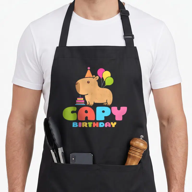 Rodent birthday apron - Cartoon brown capybara with hat, balloons, cake, and colorful CAPY BIRTHDAY text — shown on a black cotton canvas apron against a white background