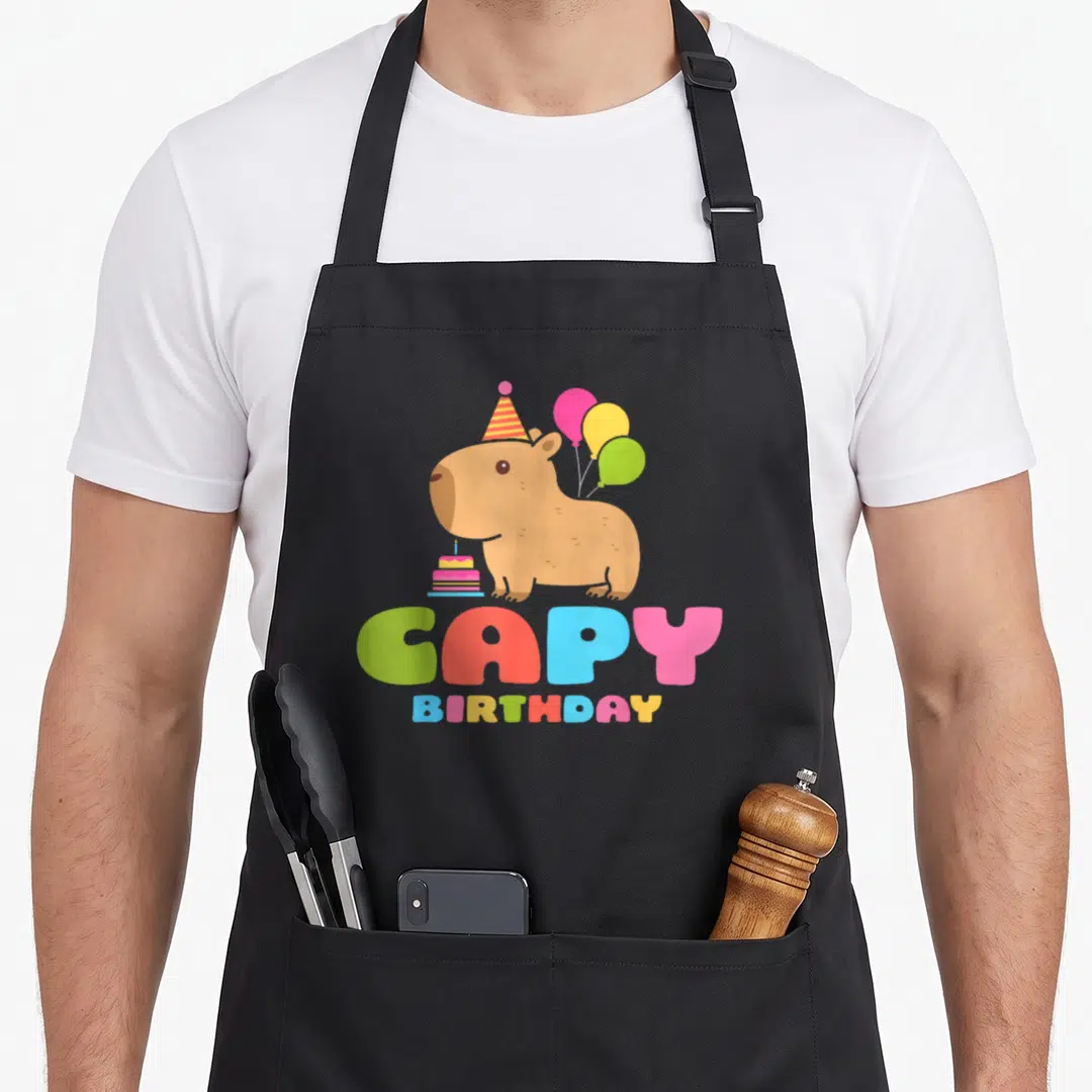 Rodent birthday apron - Cartoon brown capybara with hat, balloons, cake, and colorful CAPY BIRTHDAY text — shown on a black cotton canvas apron against a white background