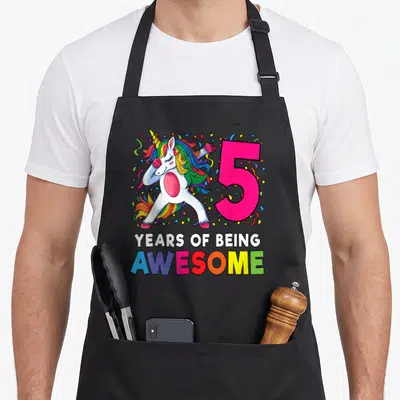 5th dabbing birthday apron - Cartoon unicorn dabbing beside pink number 5 and rainbow text celebrating five years — shown on a black cotton canvas apron against a white background