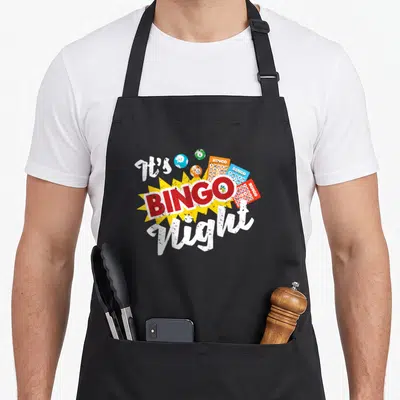 Night gambling bingo apron - It's BINGO Night text with red banner, yellow starburst, cards, and colored balls — shown on a black cotton canvas apron against a white background